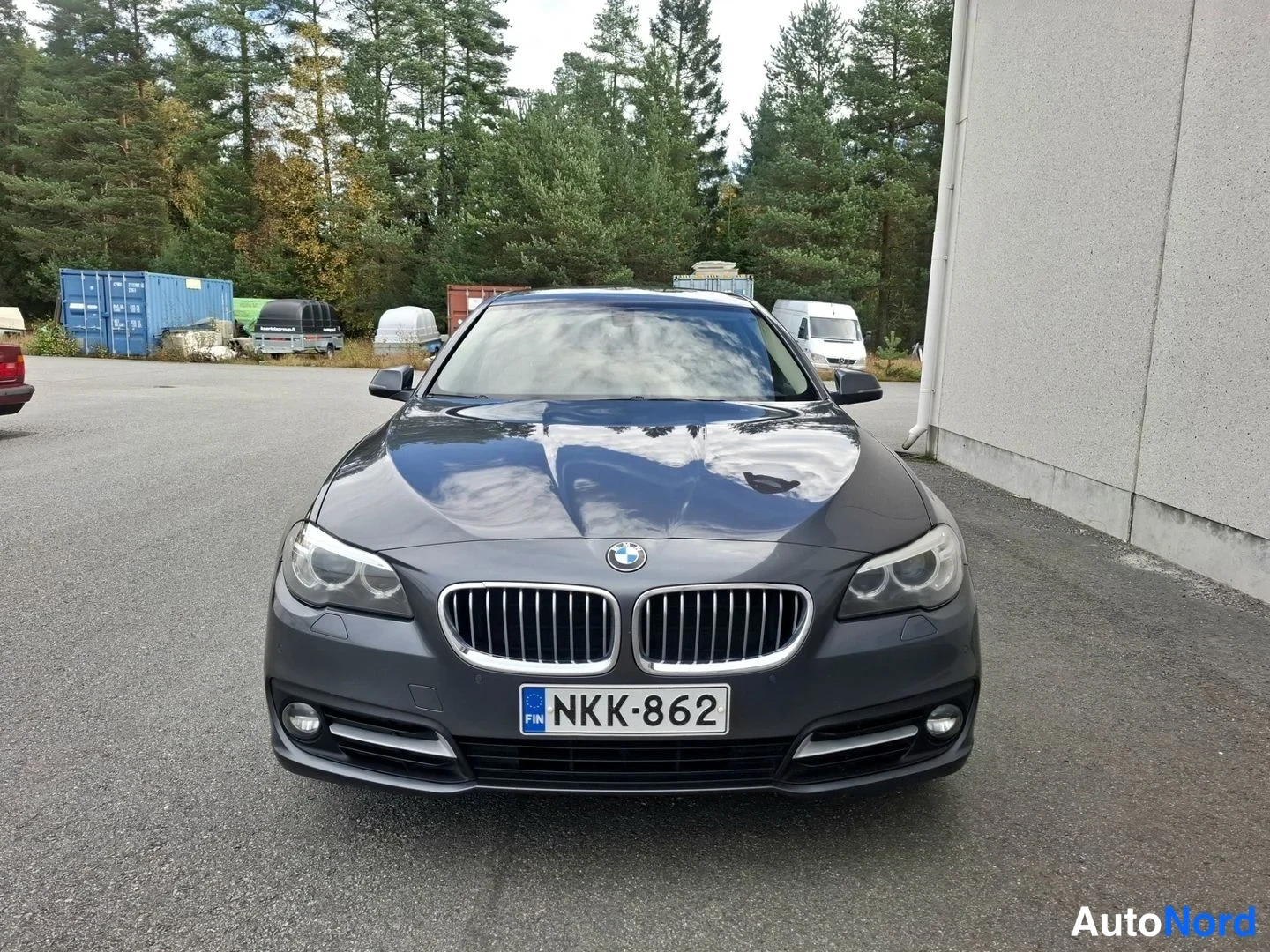 Edestä: Tummanharmaan BMW-sedanin etupää suoraan edestä kuvattuna: munuais säleikkö, ajovalot ja etupuskuri näkyvät, rekisterikilpi lukee.