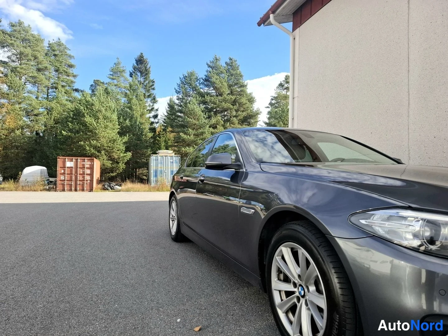 Etuviisto, nokka oikealle: Tummanharmaa BMW-sedan kolmivartalokuvakulmasta, keula ja vasen sivu näkyvällä, asfalttipihalla rakennuksen vieressä.