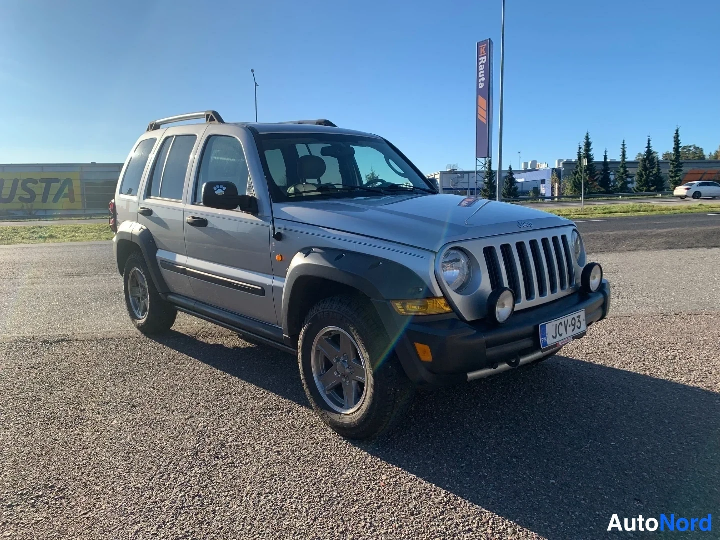 Etuviisto, nokka oikealle: Hopeanvärinen Jeep-katumaasturi kolmannesovelta edestä oikealta; korit näkyvät auringonvalossa, rekisteritunnus.