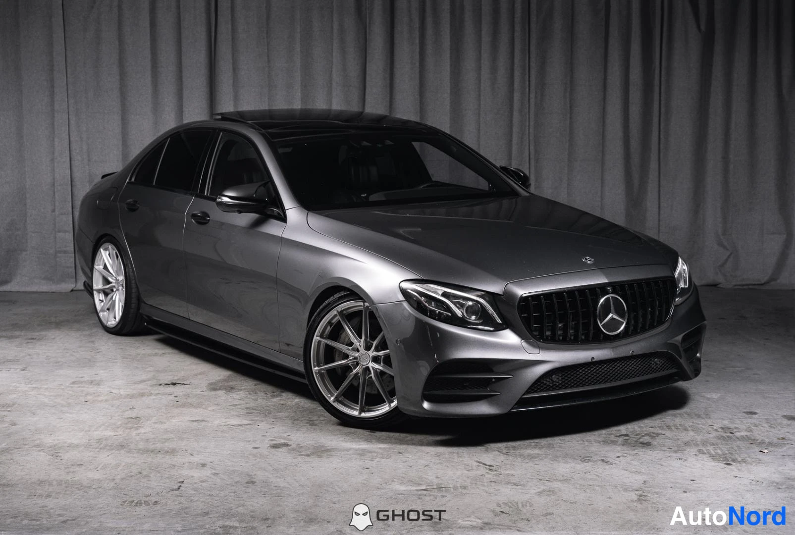 Etuviisto, nokka oikealle: Harmaa Mercedes-Benz-sedan kuvattuna etuviistosta studiossa, keula kuvan oikeassa laidassa, hopeat kevytmetallivanteet.