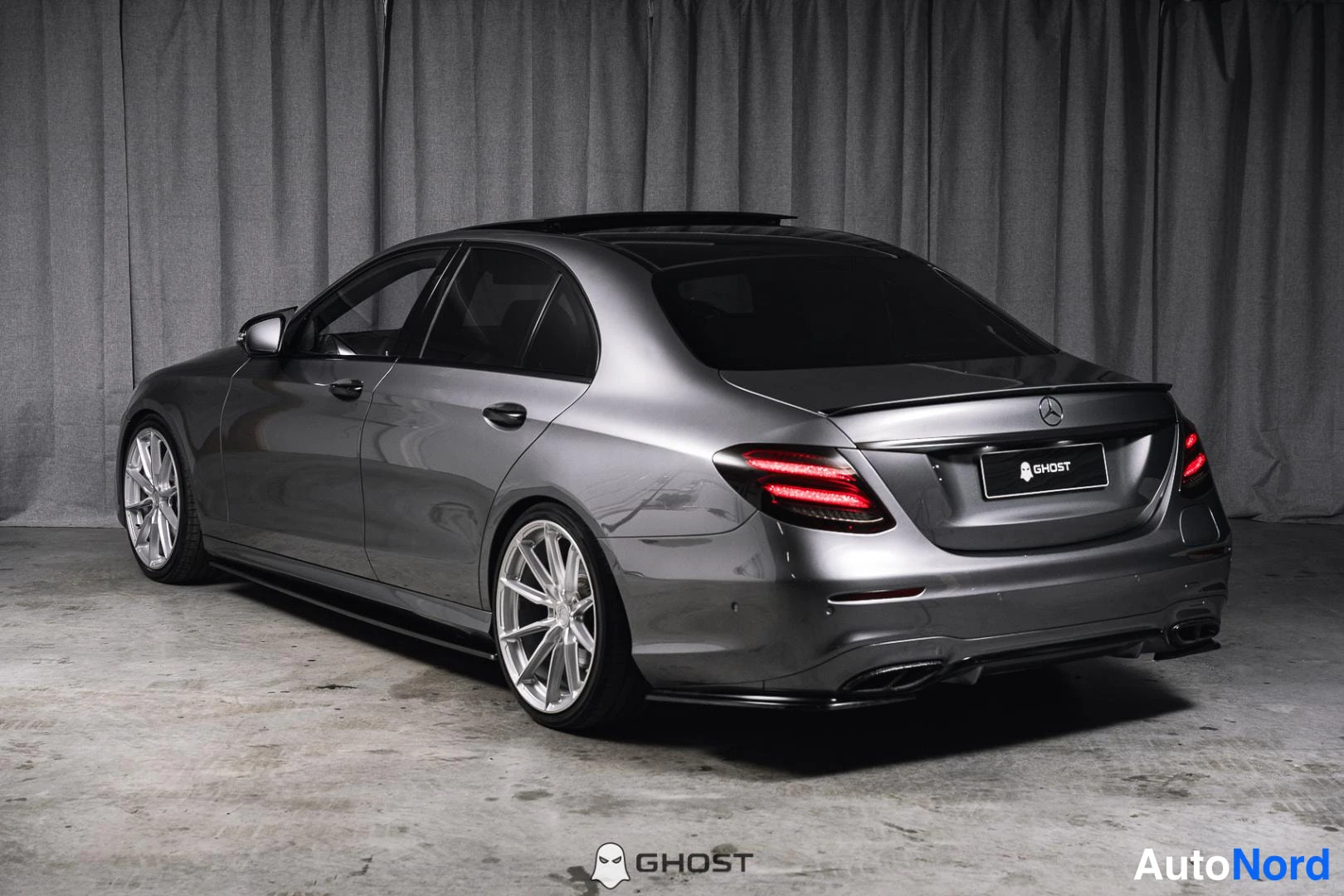 Takaviisto: Metallinvärisen Mercedes-Benz-sedan takaosa ja vasen kylki. Takavalot palavat.