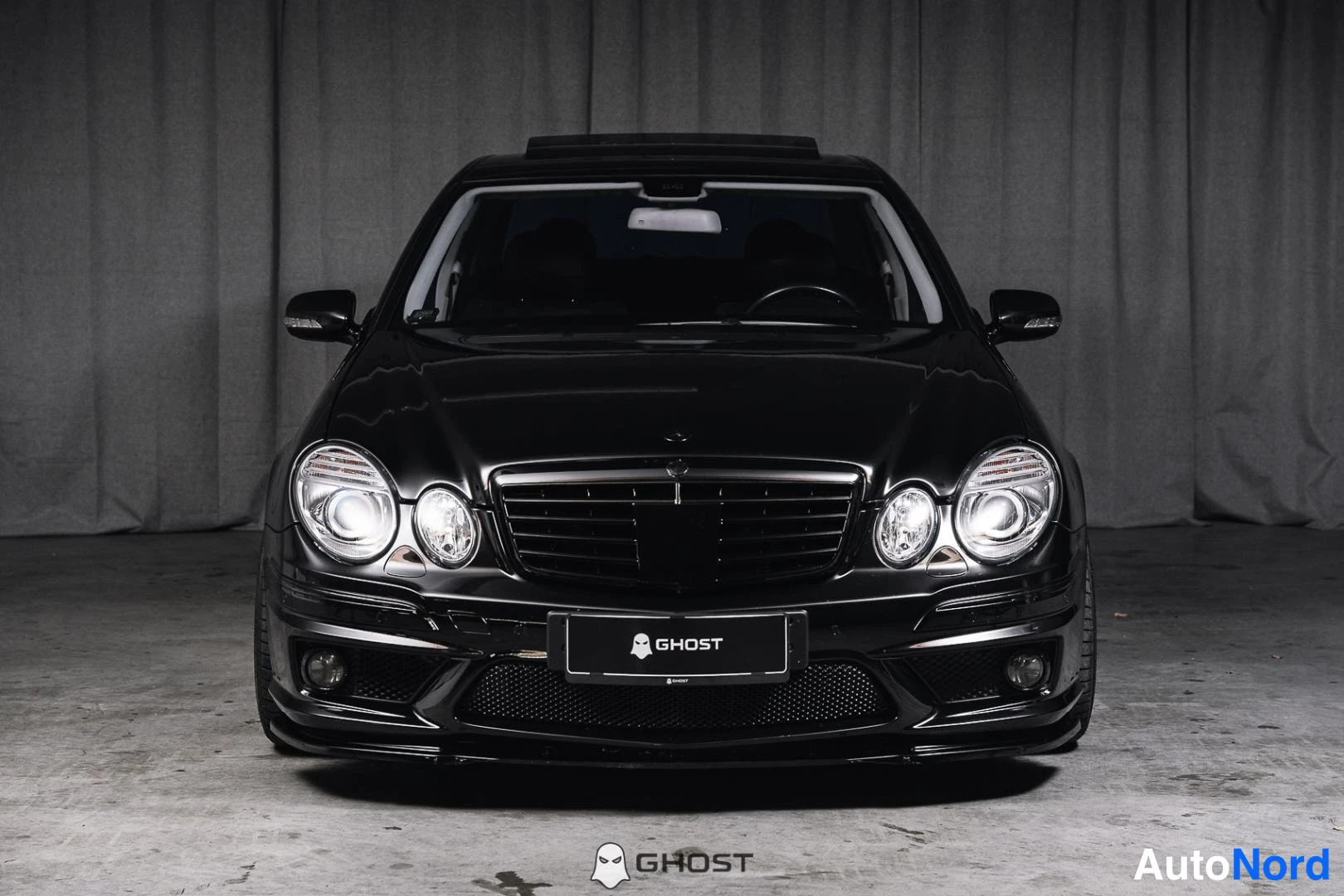 Edestä: Mustan Mercedes-Benz-sedan etunäkymä suoraan edestä, pyöreät ajovalot ja peruslainen rekisteritunnus kilpi.