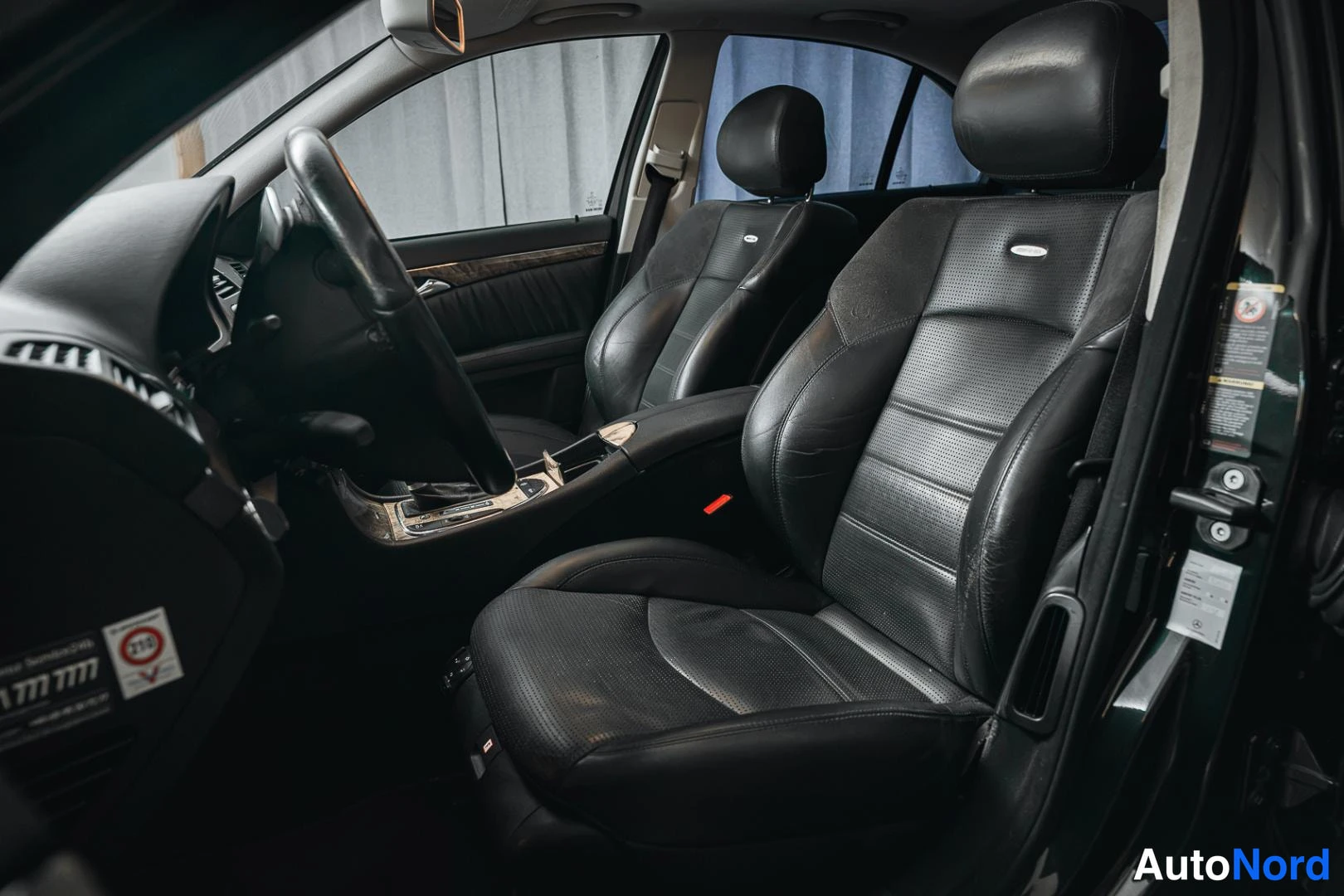 Etuistuimet: View from the driver's door frame looking into the cabin, showing two individual front bucket seats upholstered in black perforated leather and the steering whe