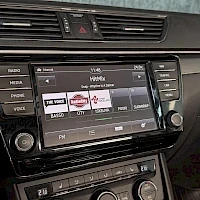 Yksityiskohta: Auton keskikonsolin mediasoitin, näytössä radiovalikko ja painikkeet kuten MENU, RADIO, MEDIA, PHONE, VOICE ja NAV. Kuva on hieman sumea ja osittain heijastuva.