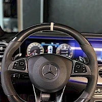 Etuistuimet: Ohjauspyörän lähikuva. Keskellä on Mercedes-logo ja ohjauspään molemmin puolin näkyvät painikkeet. Mittaritaulun sininen taustavalo näkyy.