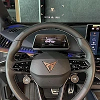 Yksityiskohta: Close-up of the steering wheel with digital instrument cluster visible behind it. The Dacia logo is in the center of the wheel.