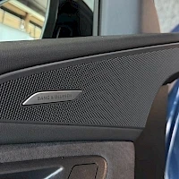 Yksityiskohta: Detail shot of a door panel speaker grille with a chrome trim piece embossed with the text 'BANG & OLUFSEN'.