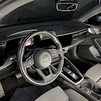 Etuistuimet: Black leather-wrapped multifunction steering wheel, dashboard with digital display, and light-colored seats of the driver's side.