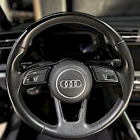 Yksityiskohta: Close-up of a black Audi multifunction steering wheel featuring silver control buttons on both sides and the four-ring logo.