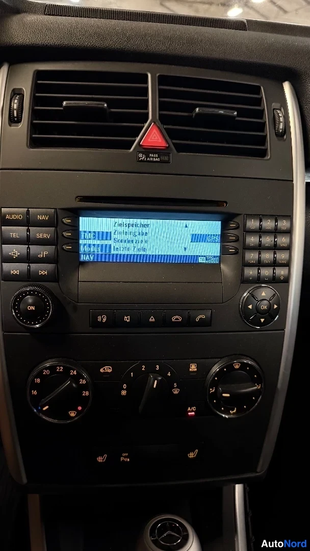 Yksityiskohta: Mercedes-Benzin kojelaudan keskiosa: radio- ja ilmastointipaneeli, näytöllä radiokuuntelu ja kielivalikko, alapuolella lämpötilan säätökiekot ja A/C-nappi.