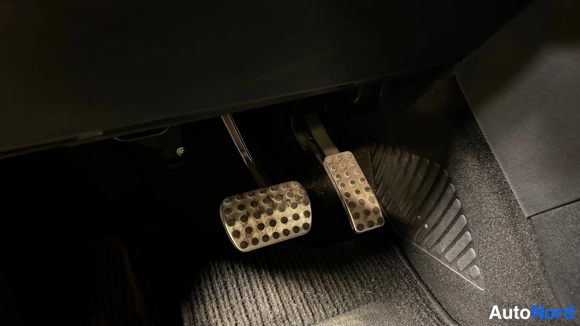 Yksityiskohta: Lähikuva autoon kuuluvista poljinlaudoista (kaasupoljin ja jarrupoljin), metallipinnassa olevat pyöreät irrotuspisteet. Poljinten alla tumma matto.