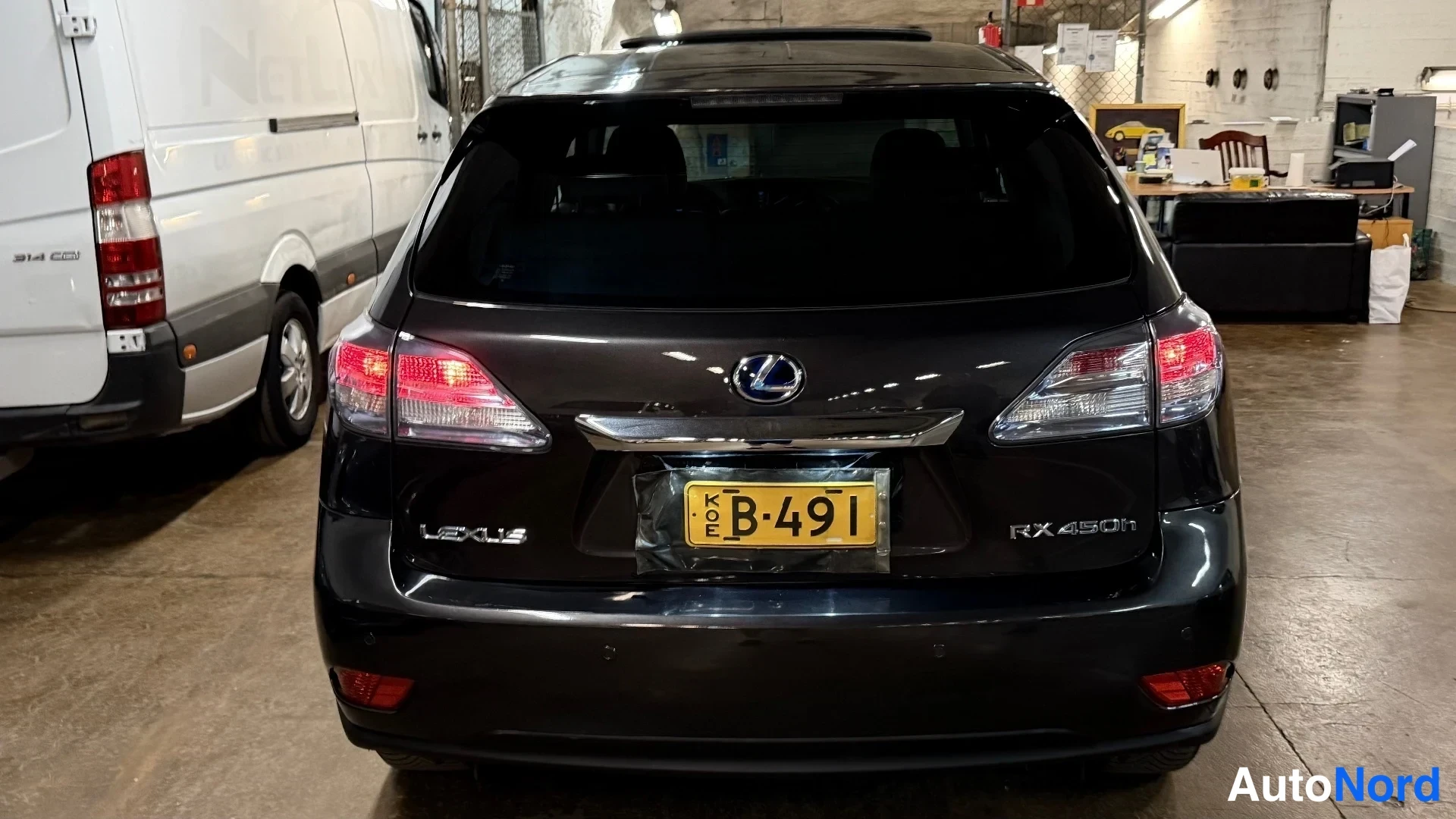 Takaa: Täysin kuvattu mustan Lexus RX450h:n takapää sisätiloissa. Keskellä keltainen autonumerokilpi B-491 (KO E), kyljessä 'LEXUS' ja oikealla 'RX450h'. Takalamppujen
