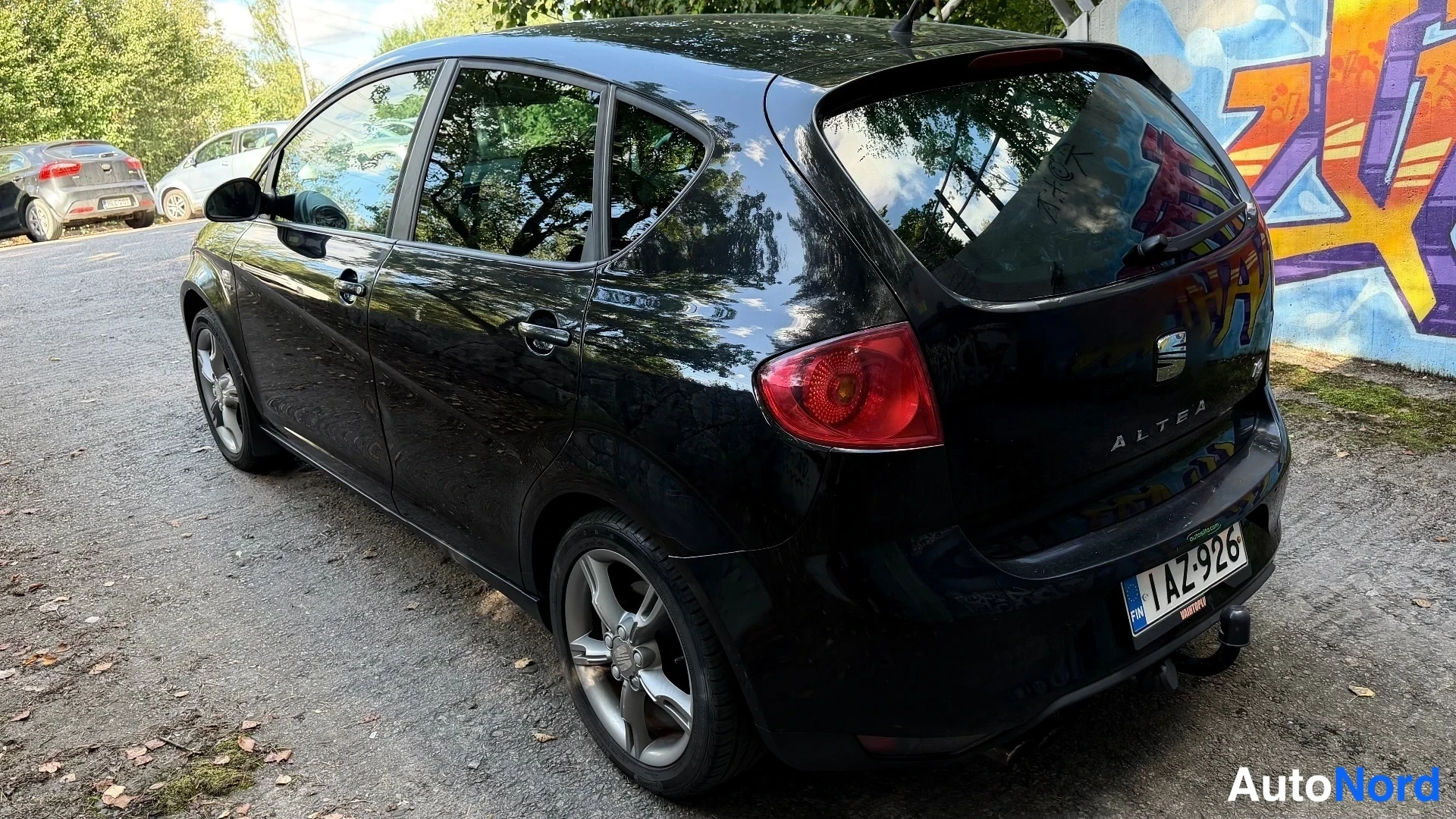 Takaviisto: Mustan Seat Altean takakulma. Rekisterikilpi, takaluukussa ALTEA-teksti ja Seat-logo, takakoukku asennettu. Auto seisoo sorapohjaisella parkkipaikalla graffitii