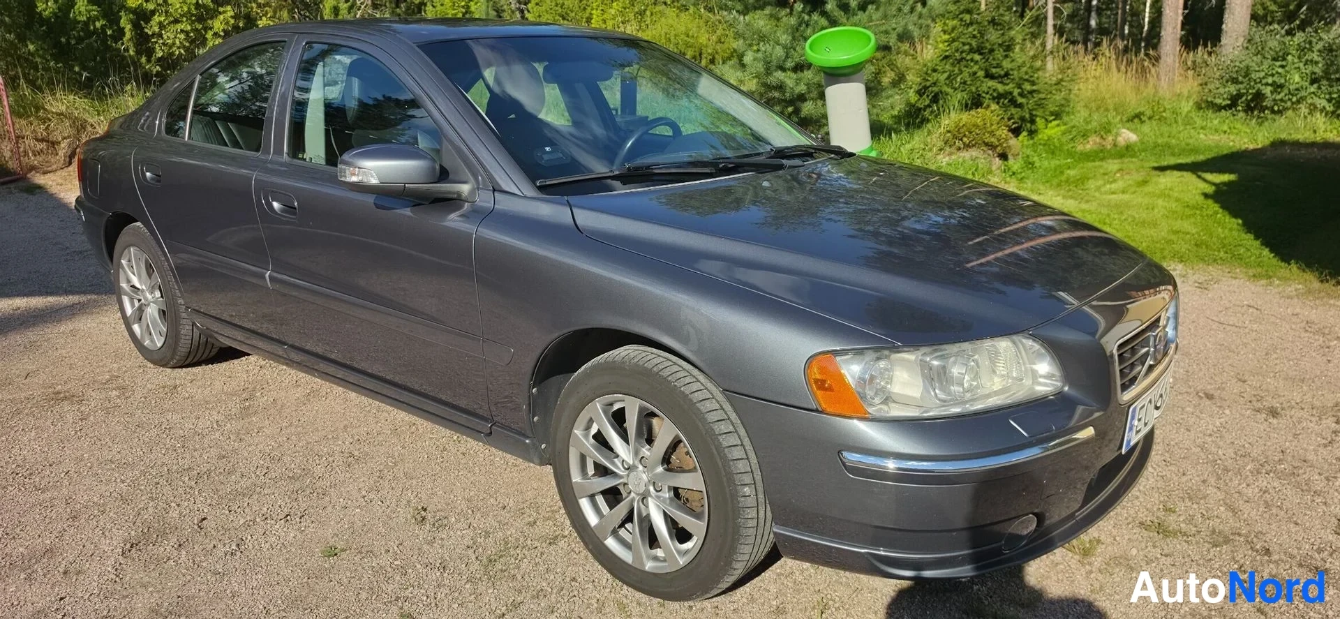Etuviisto, nokka oikealle: Tummanharmaa Volvo S60 etuviistosta oikealle kuvattuna sorapohjaisella pihalla, vihreä esiintymä taustalla.