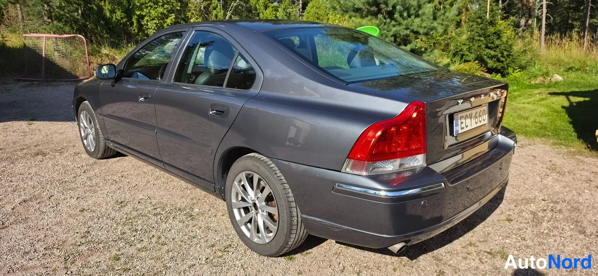 Takaviisto: Tummanharmaa Volvo S60 takaovistosta oikealta kuvattuna, rekisterikilpi, sorapohjainen piha ja metsä taustalla.
