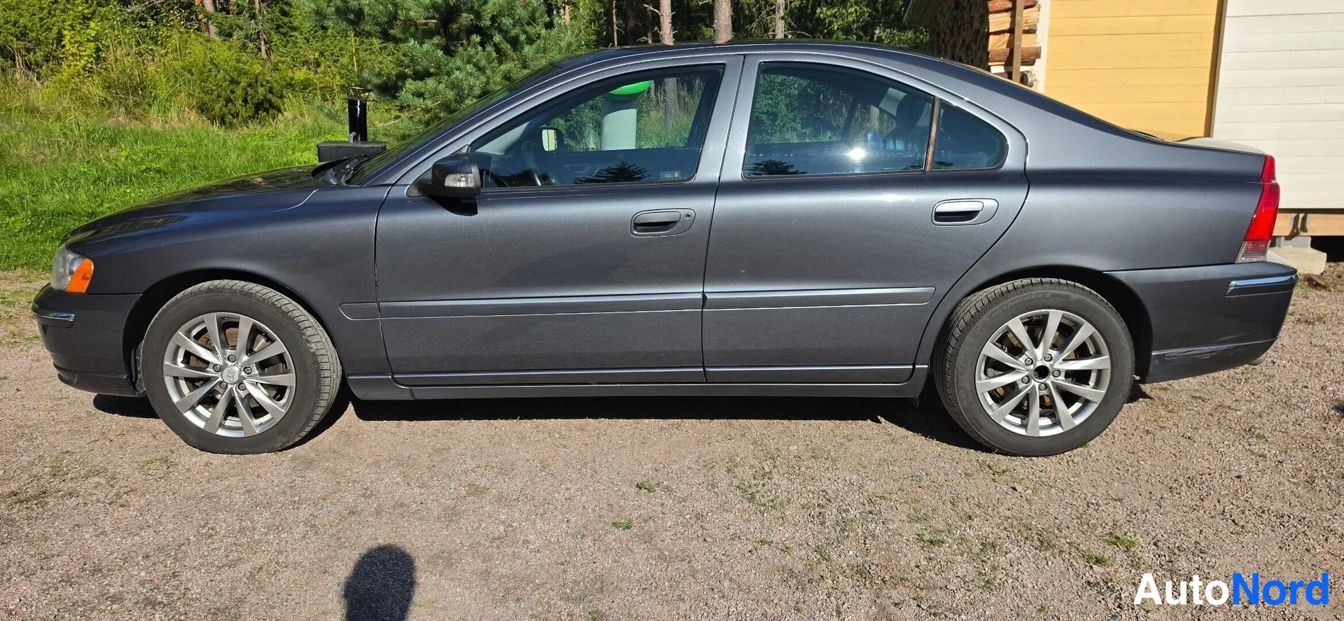 Etuviisto, nokka vasemmalle: Volvo S60 -sedan sivulta-etuvinkkelistä vasemmalla, tummanharmaa, talvella/dirt-free hiekkamaalla rakennusten edessä.