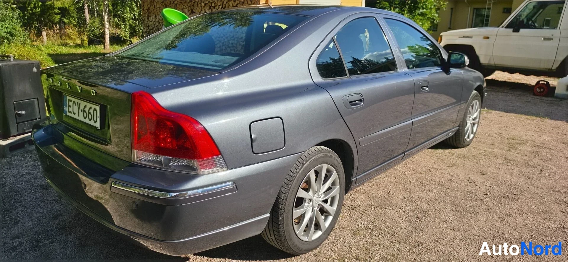 Takaviisto: Volvo S60 takaosa vinosta oikealta, tummanharmaa, rekisterikilpi, takana hirsipino ja pakettiauto.