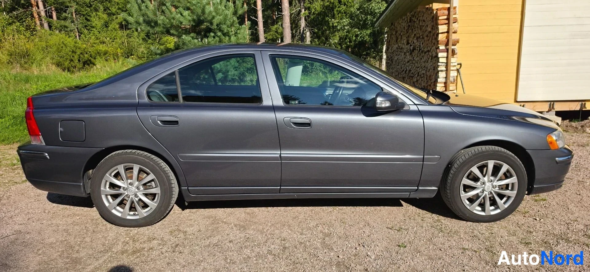 Etuviisto, nokka oikealle: Volvo S60 -sedan vasen kylki ja keula-osittain näkyvissä oikealla, tummanharmaa, hiekkapinta ja keltainen rakennus taustalla.