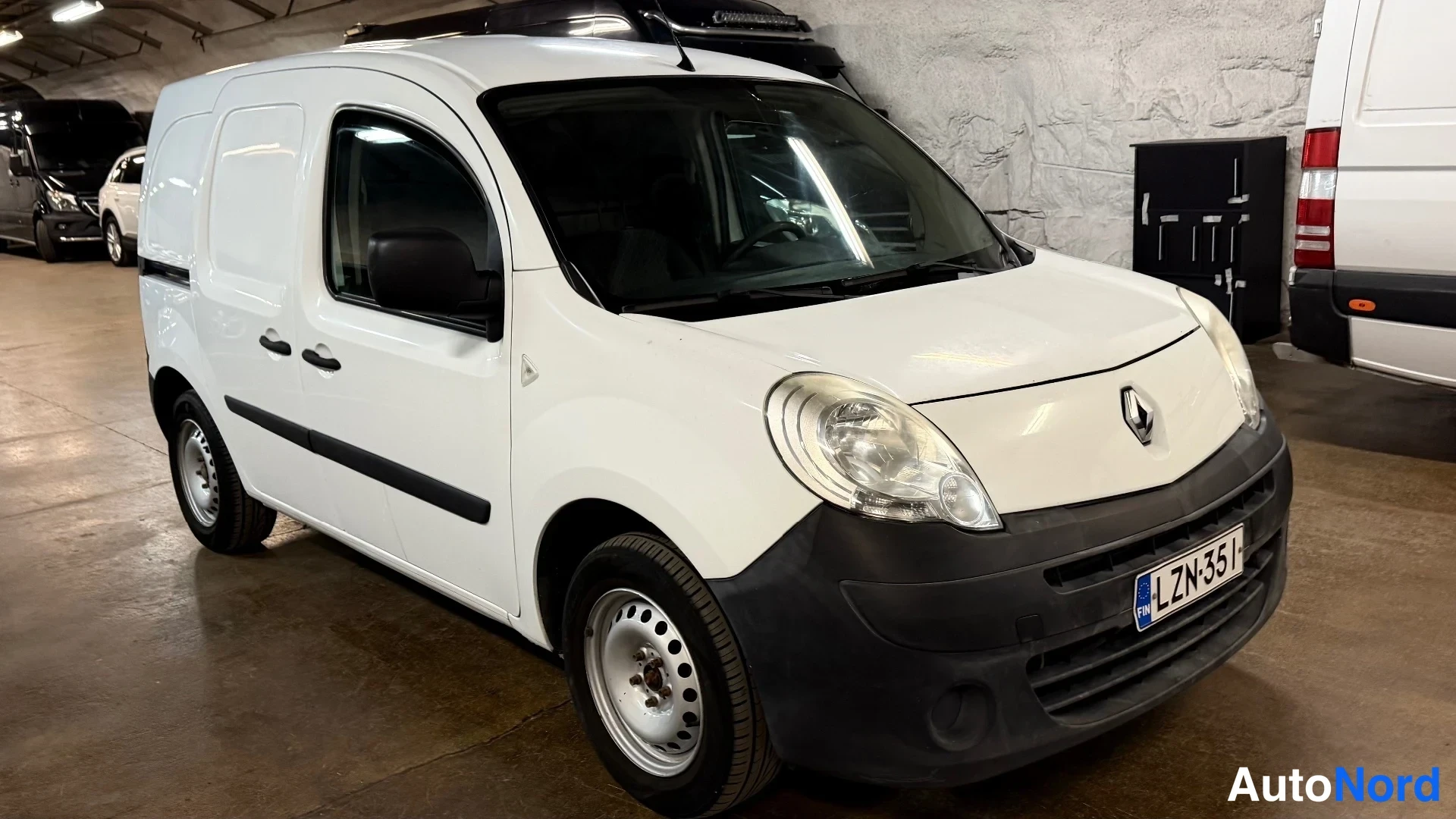 Etuviisto, nokka oikealle: Valkoinen Renault Kangoo -pakettiauto etukulmasta oikealle kuvattuna parkkihallissa, rekisterikilpi.