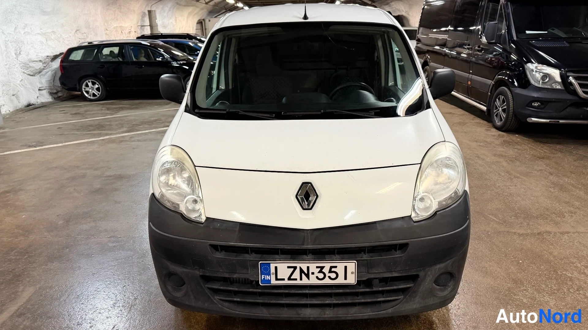 Edestä: Valkoisen Renault Kangoo -pakettiauton etunäkymä suoraan edestä parkkihallissa, rekisterikilpi.
