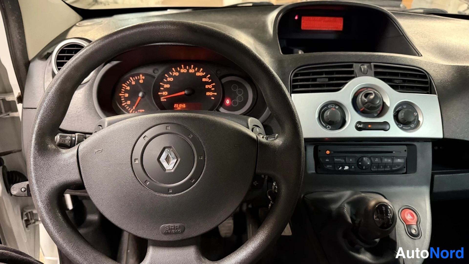 Muu: Ohjaamon näkymä: ratissa Renault-merkki, takana oranssit taustavalaistut mittarit ja keskikonsolilla ilmastointisäätimet ja radio.