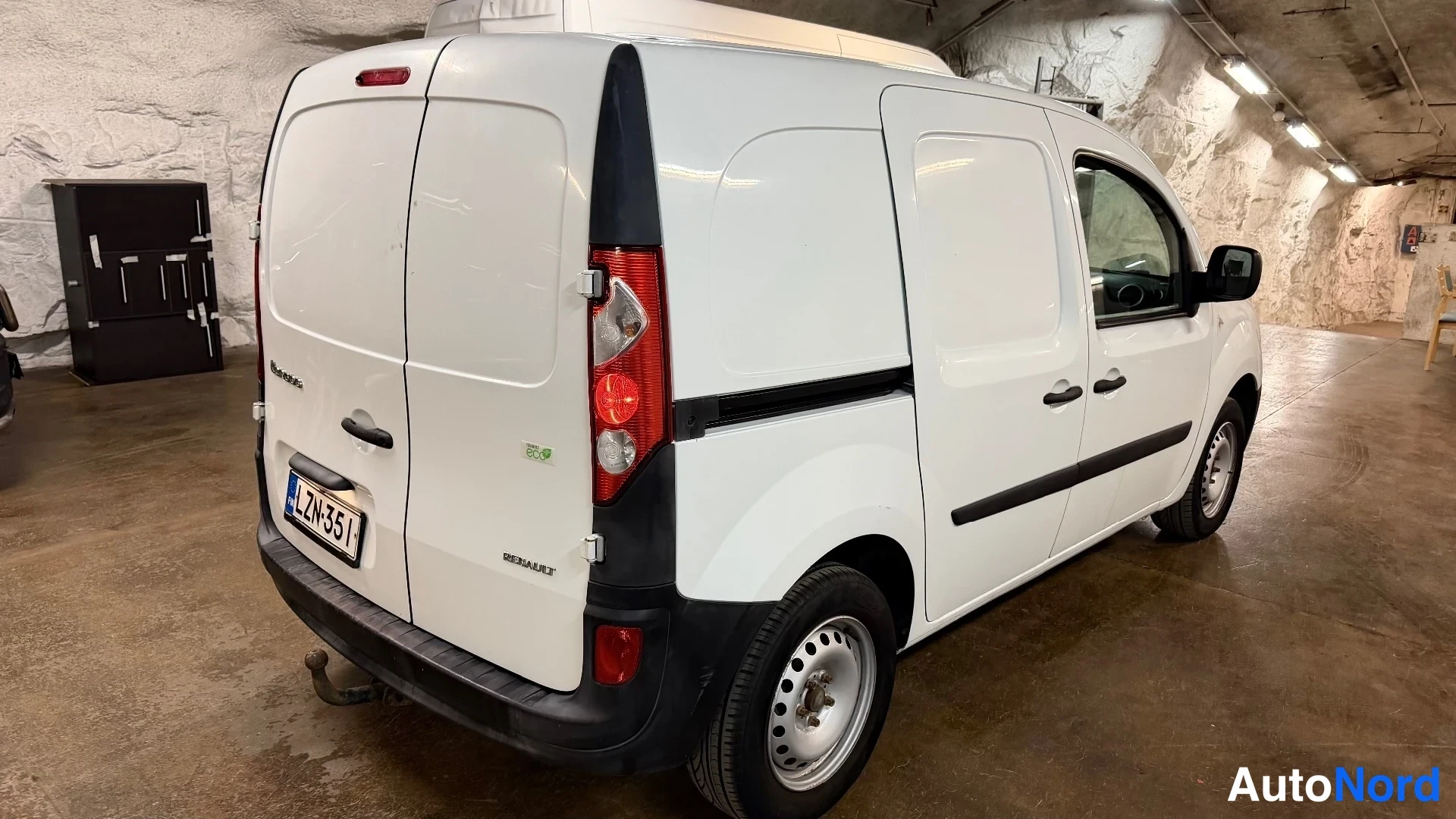 Takaviisto: Rear three-quarter view of a white Renault Kangoo Express van in a garage; rear face (boot lid, taillights, 'RENAULT' badge), right flank, and license plate vis
