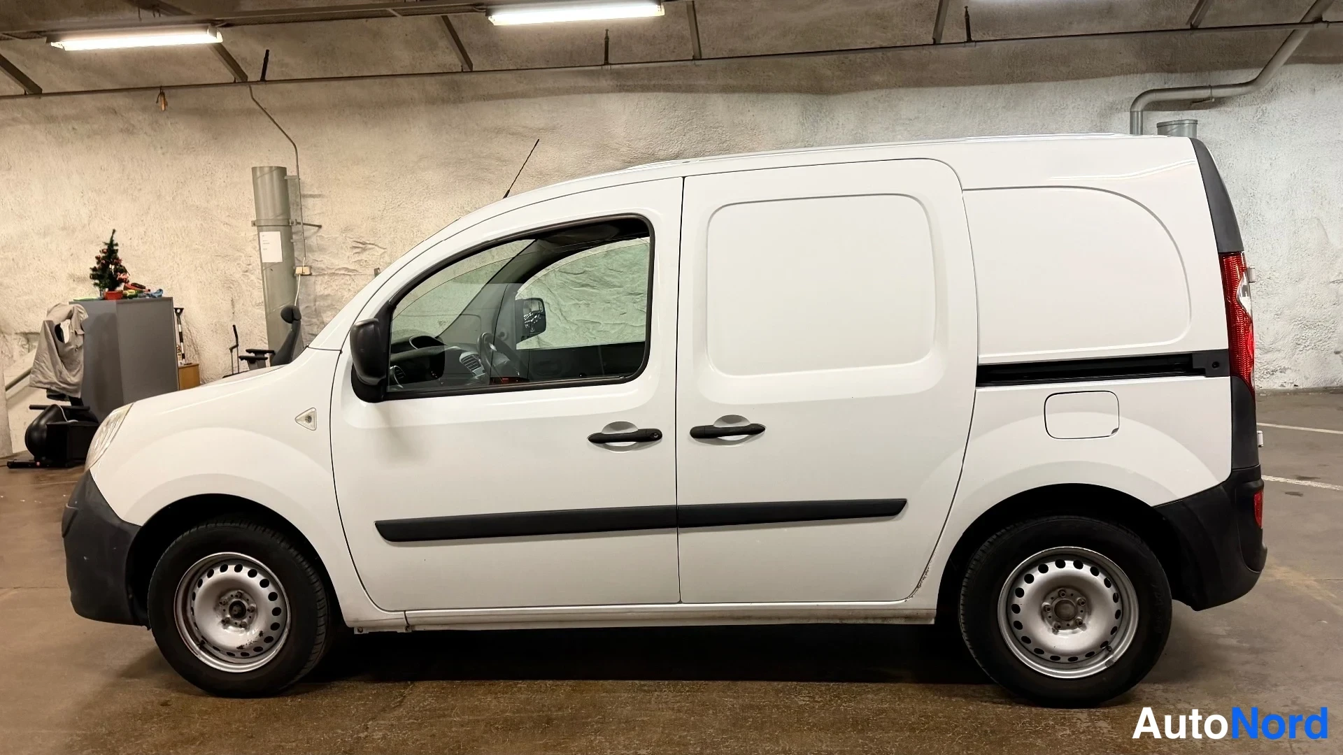 Sivulta: Side profile of a white Renault Kangoo Express van in a garage, showing the right flank, passenger and cargo door windows, front headlight, black side trim, ste