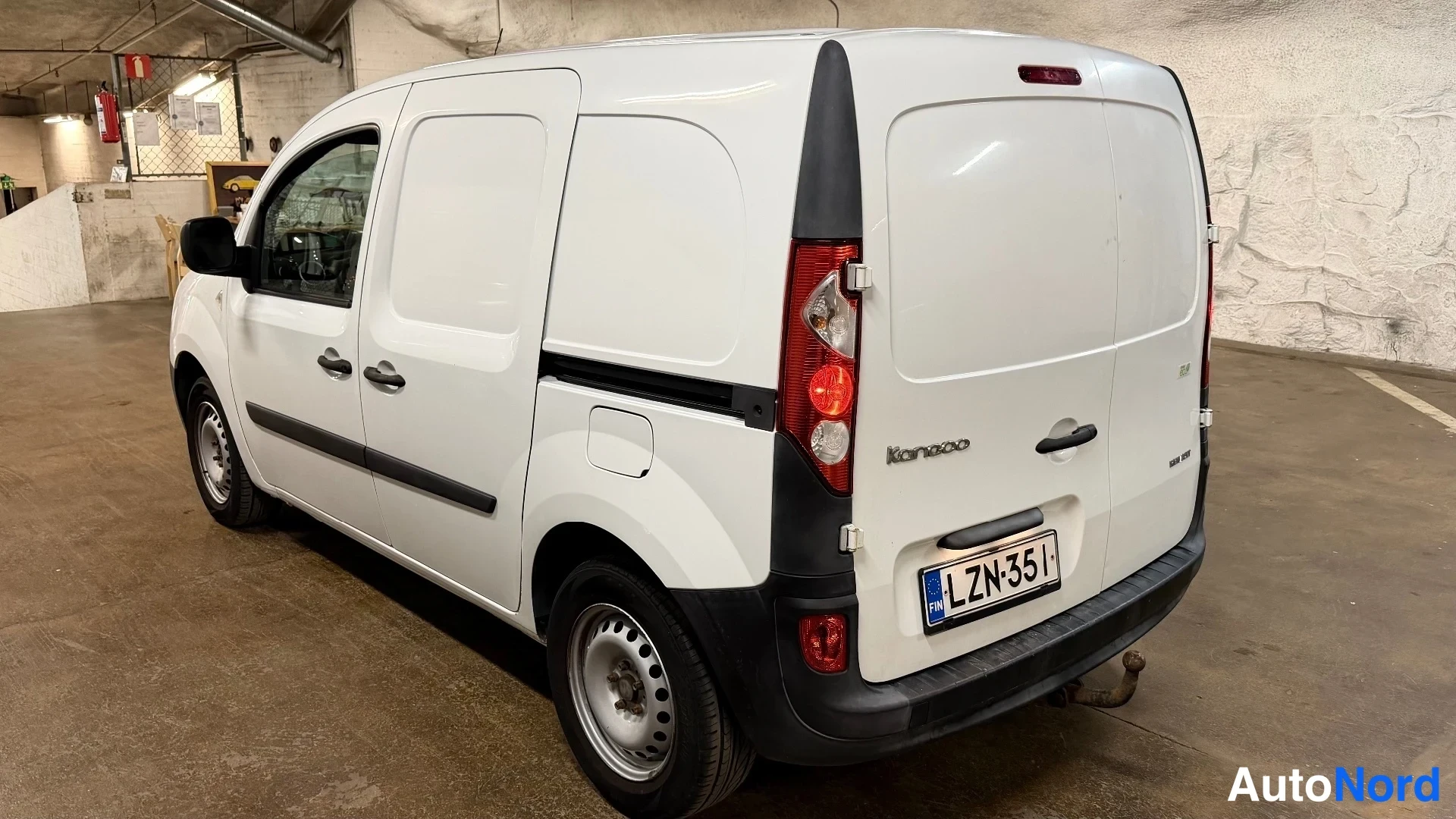 Takaviisto: Takaa ja oikealta puolelta otettu kuva valkoisesta Renault Kangoo -pakettiautosta sisäparkkipaikalla. Takaluukussa lukee KANGOO. Autossa on perävaunu koukku.