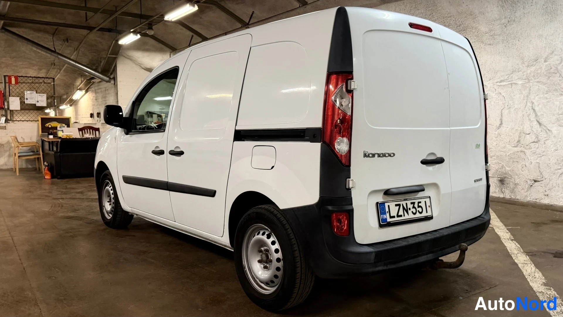 Takaviisto: Takaa ja oikealta puolelta otettu leveä kuvakulma valkoisesta Renault Kangoo -pakettiautosta sisätilassa. Auton takana on työhuoneen näköinen alue tuoleineen. A