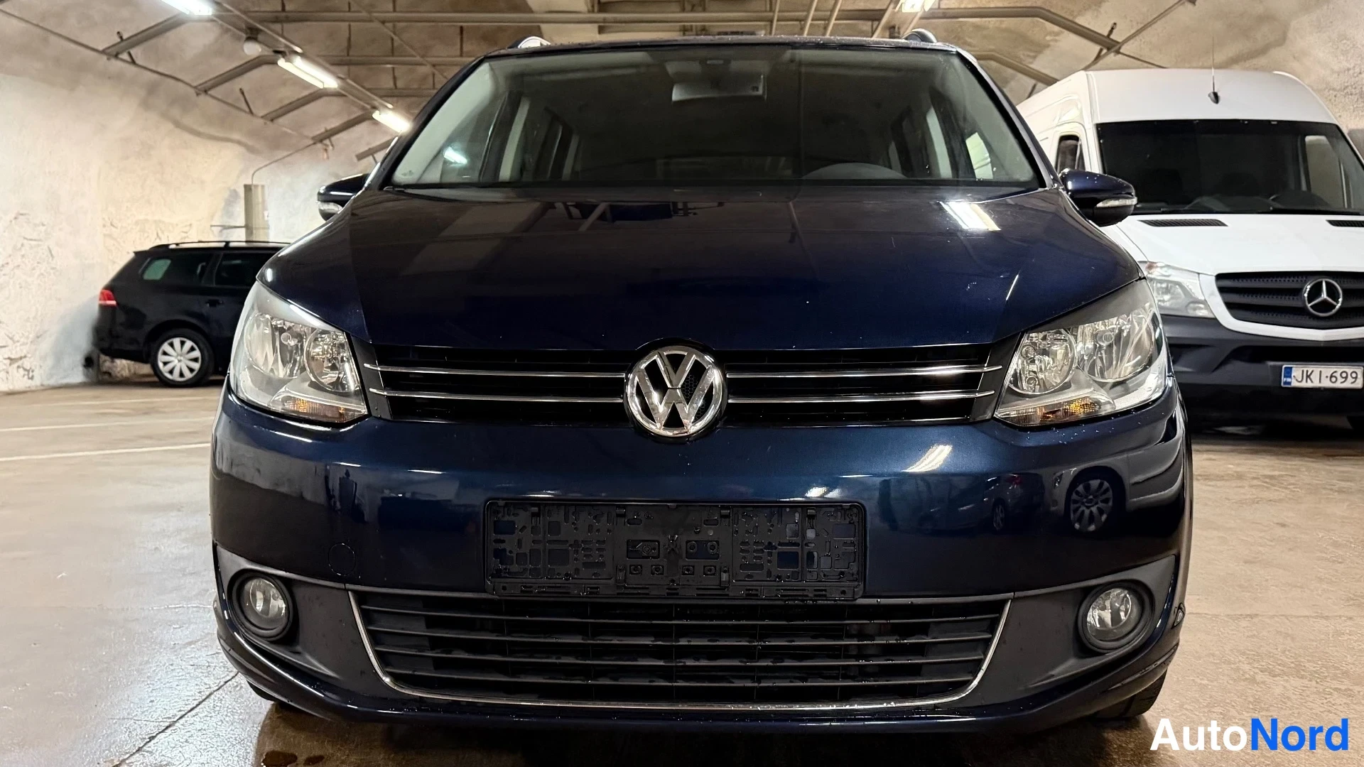Edestä: Straight-on frontal view of a dark blue Volkswagen showing the chrome grille, central badge, headlights, and blanked-out license plate area.