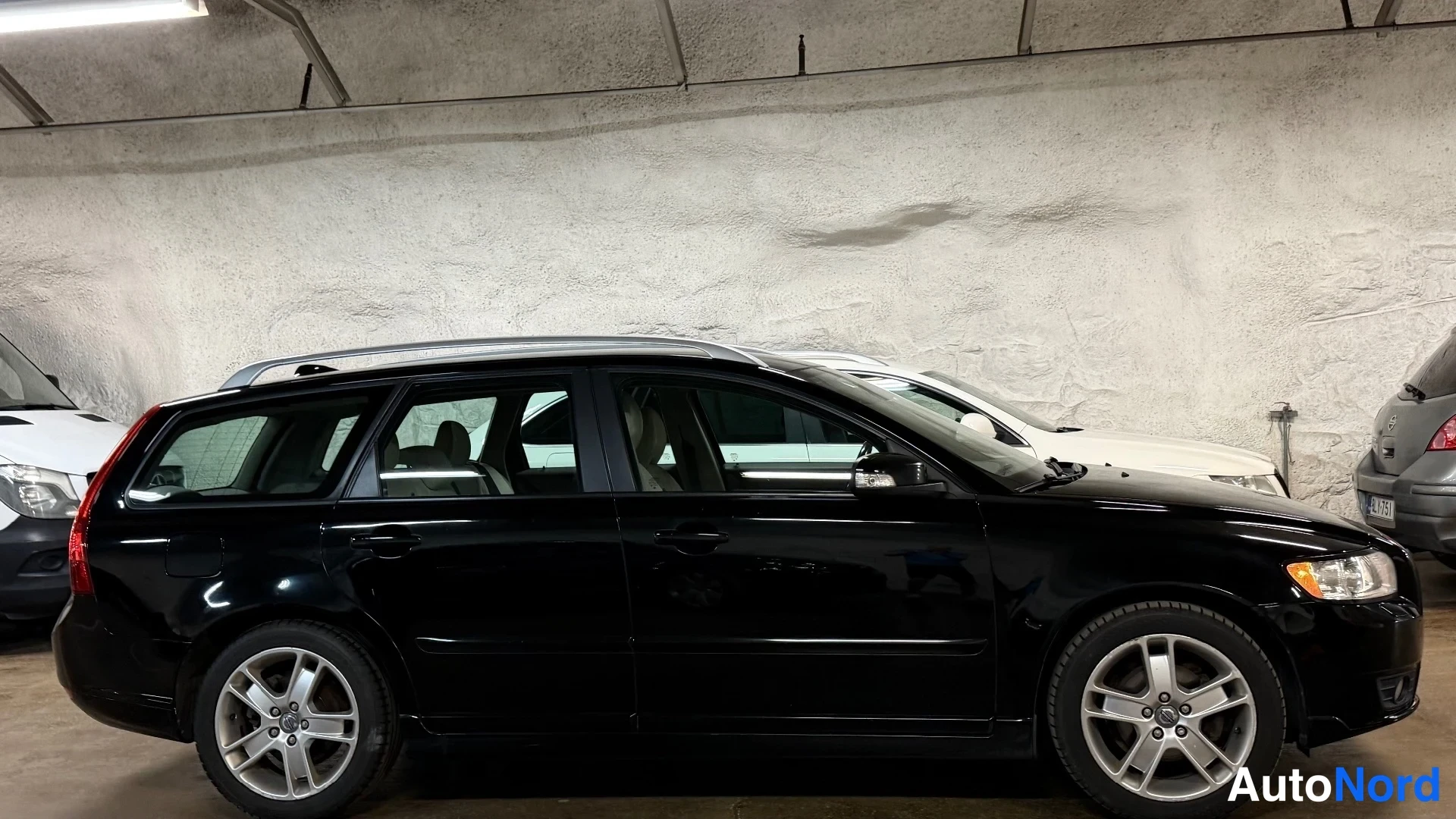 Sivulta: Musta Volvo V50 farmari seisomassa sisätiloissa; sivunäkymä, hopeat vanteet ja kattoharjat näkyvät.
