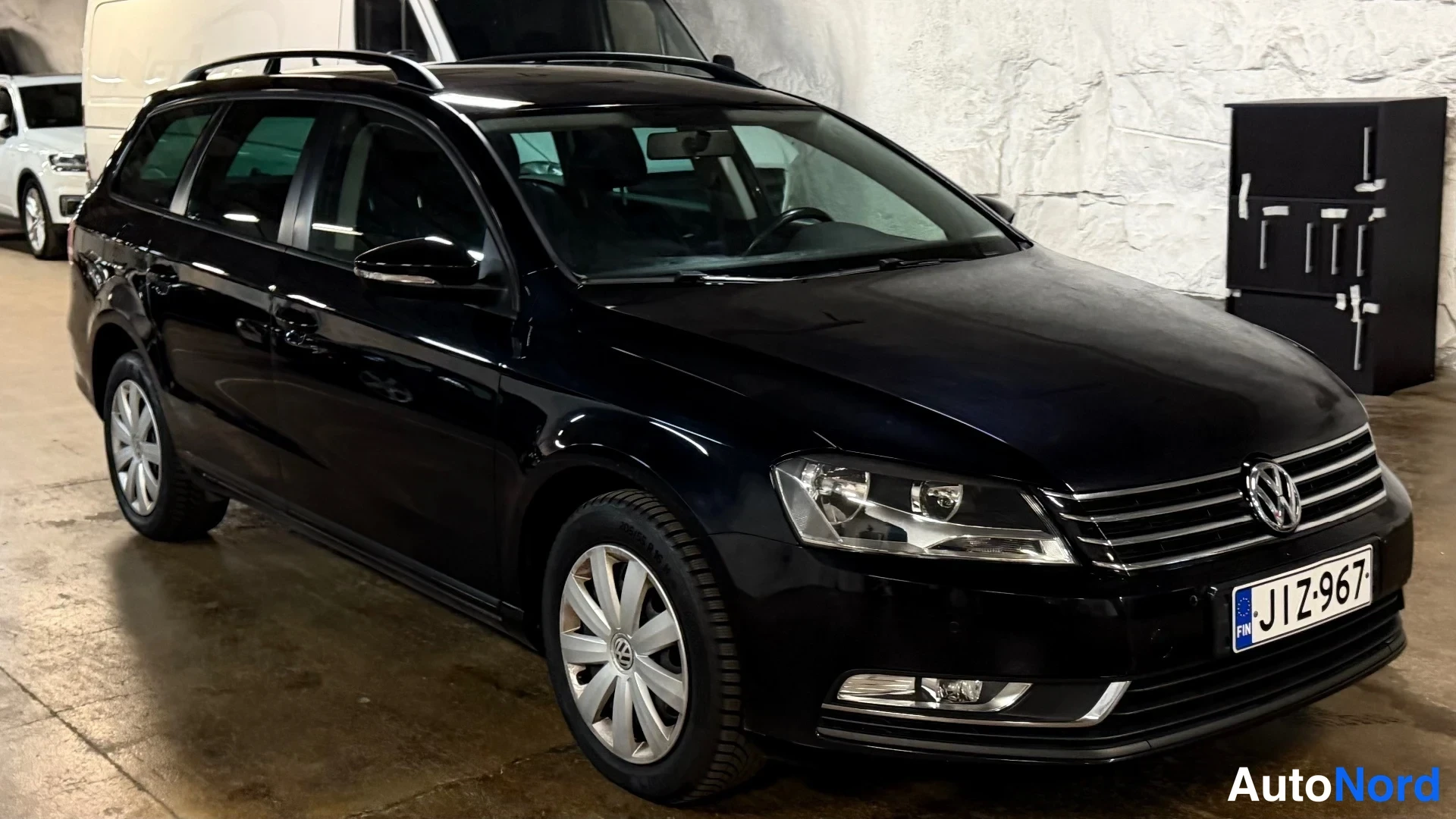 Etuviisto, nokka oikealle: Mustan Volkswagen Passat Variant -farmariauto edestä vasemmalta kuvattuna parkkihallissa, rekisterikilpi.