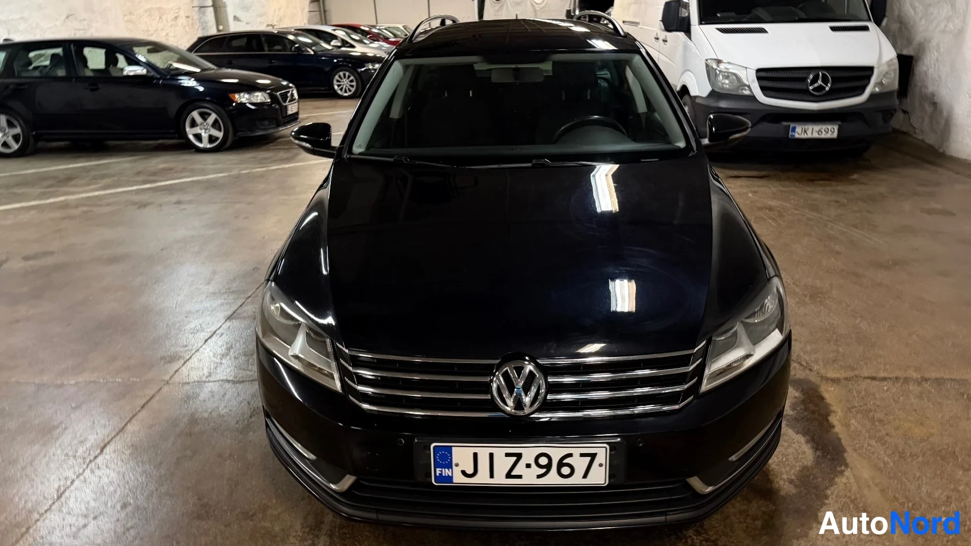 Edestä: Mustan auton etupää suoraan edestä kuvattuna parkkihallissa, rekisterikilpi.