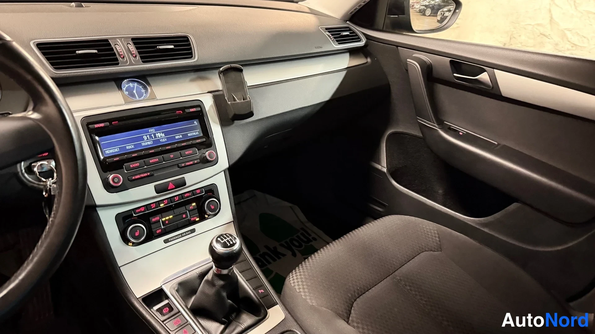 Etuistuimet: Interior dashboard close-up showing infotainment screen tuned to radio, manual gear shifter and front passenger seat.