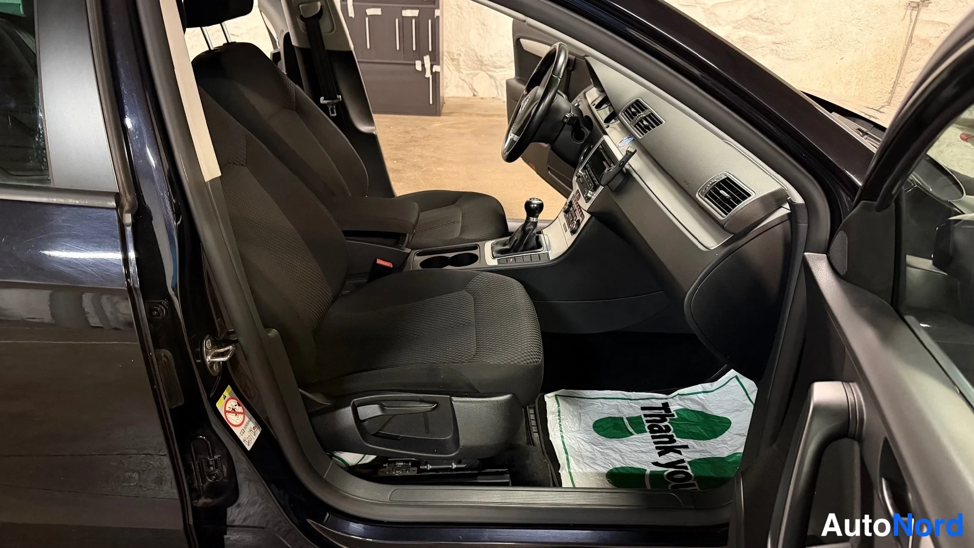 Etuistuimet: Front cabin interior viewed from open driver's side door showing two black fabric seats, dashboard and manual gear shift.
