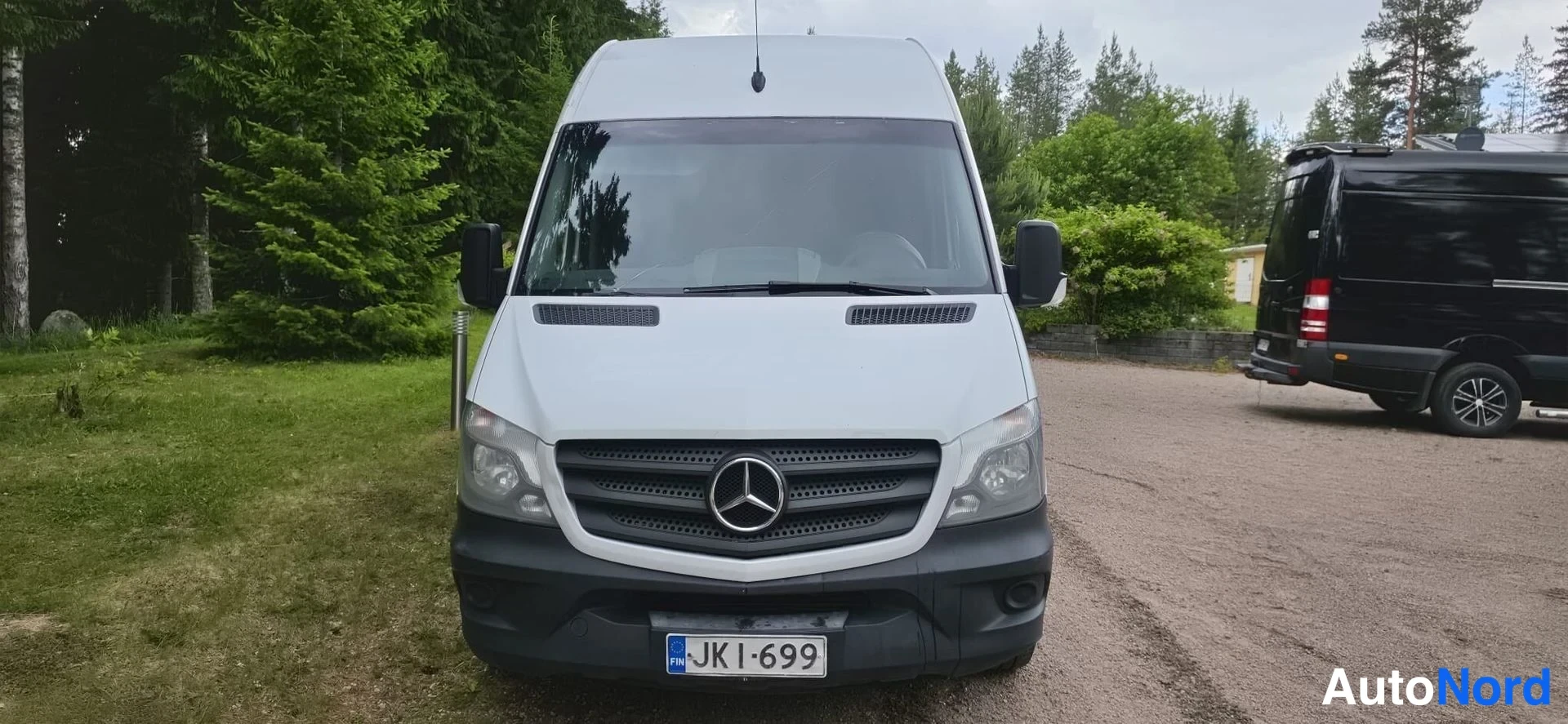 Edestä: Mercedes-Benz Sprinter -pakettiauto suoraan edestä kuvattuna; valkoinen konepelti, musta etusäleikkö Merkced-tähdellä, ajovalot ja rekisterikilpi JK I-699. Taus