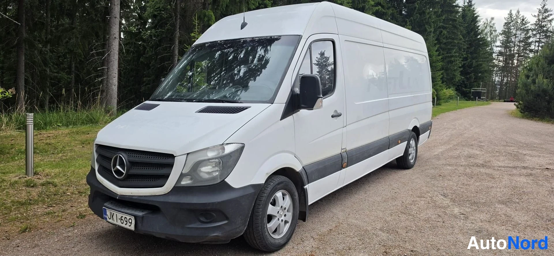 Etuviisto, nokka vasemmalle: Valkoinen Mercedes-Benz Sprinter -pakettiauto kolmivartalo-otteesta edestä vasemmalta hiekkatiellä; konepelti, musta etusäleikkö Merkced-tähdellä, ajov