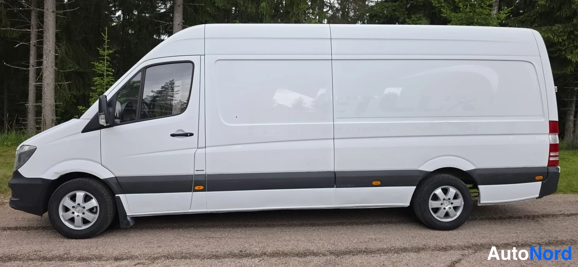 Sivulta: White van viewed from the left side. Both front wheels are visible. Sliding door tracks and black protective cladding along the bottom of the body. Trees in bac