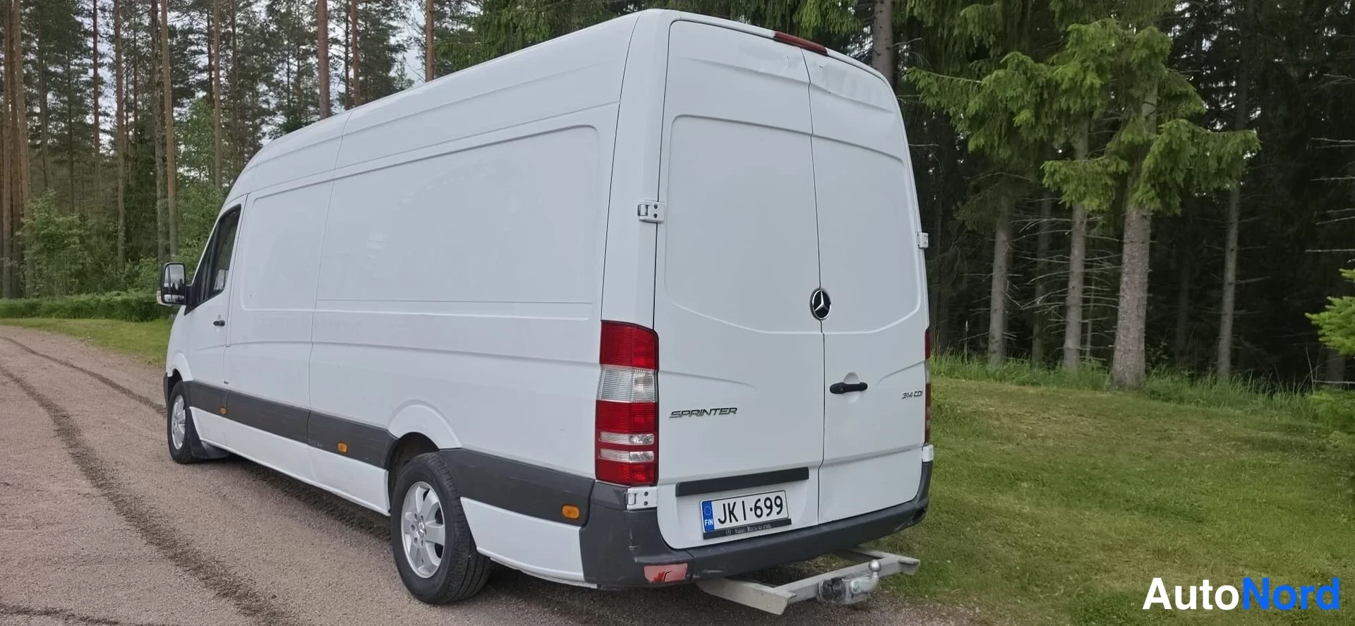 Takaviisto: White van rear view with a partial view of the right flank. Rear bumper, tail lights and license plate JK I-699 visible. Tow hitch fitted below the bumper.