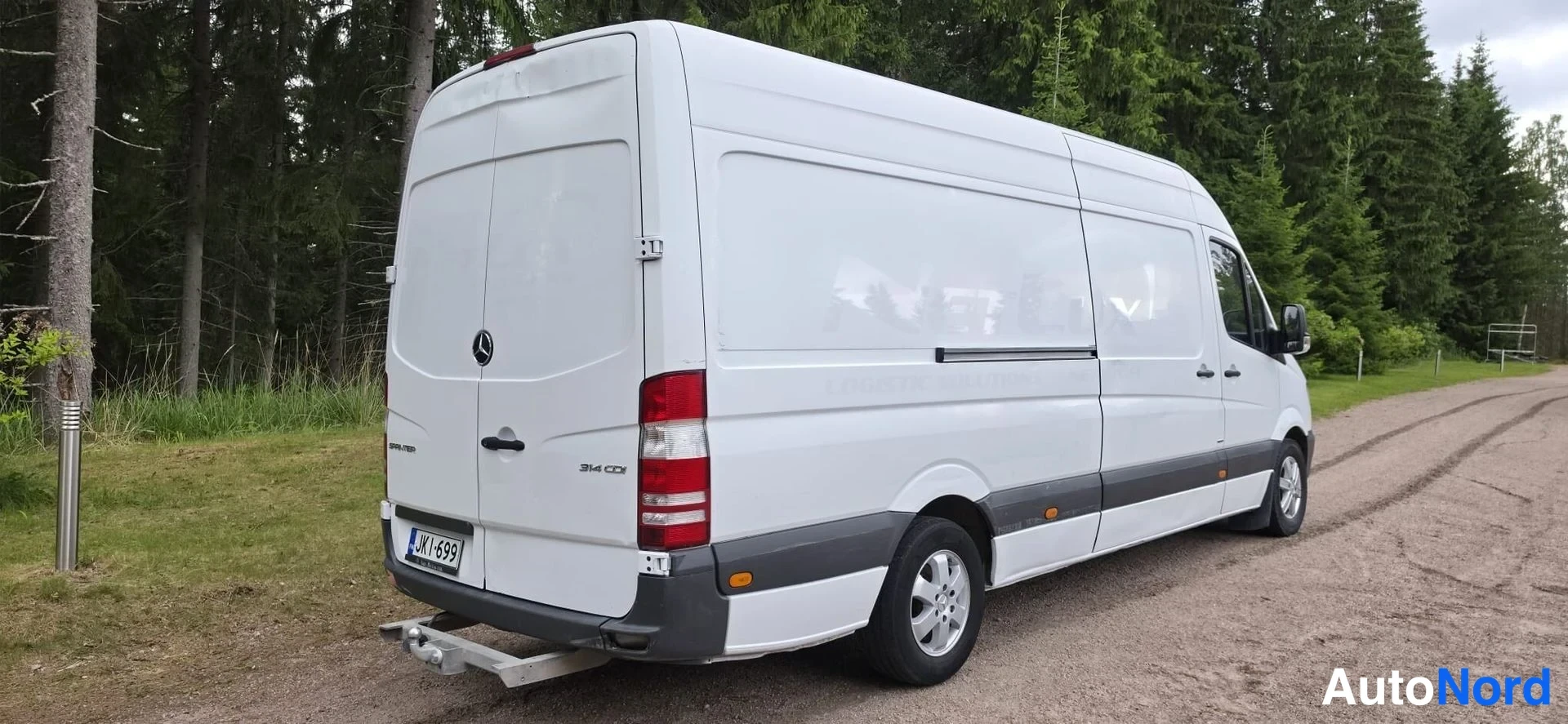 Takaviisto: White van rear view with a partial view of the left flank. Tail lights and license plate JK I-699 visible. Tow hitch fitted below the bumper.