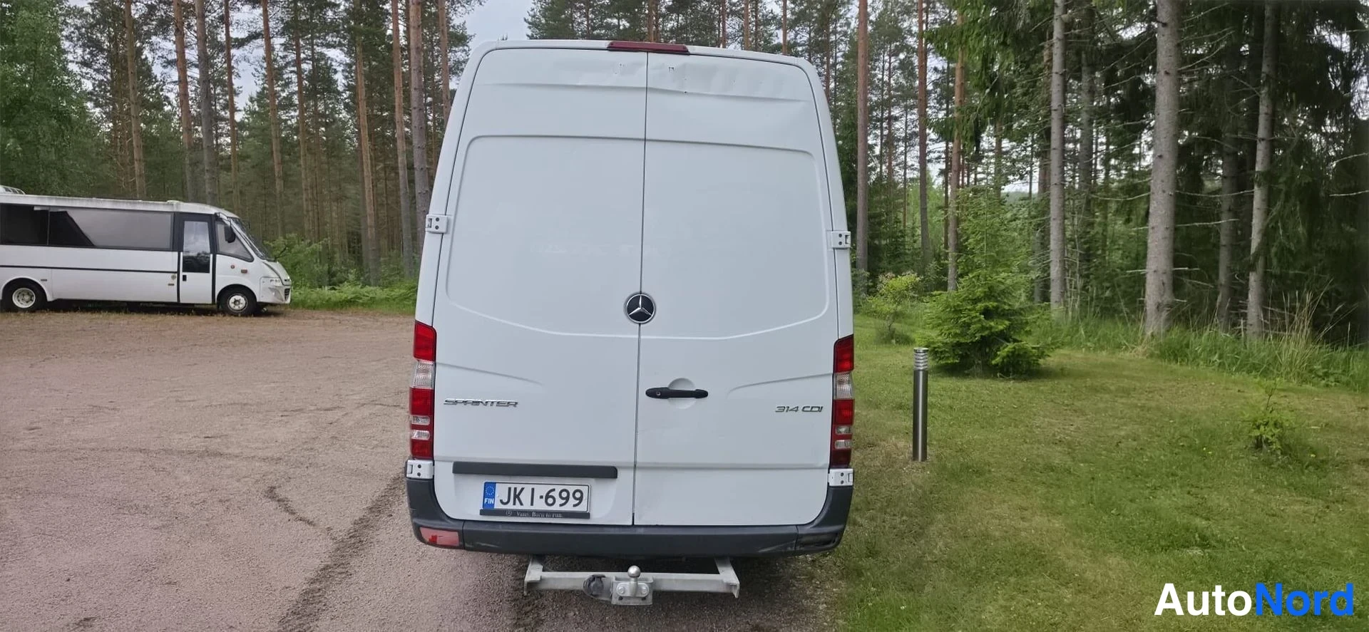 Takaa: Mercedes-Benz Sprinter -pakettiauto takaa kuvattuna, valkoinen kori, kaksiosaiset takaportit kiinni, rekisterikilpi JK I-699 ja vetokoulu; metsäinen ympäristö.