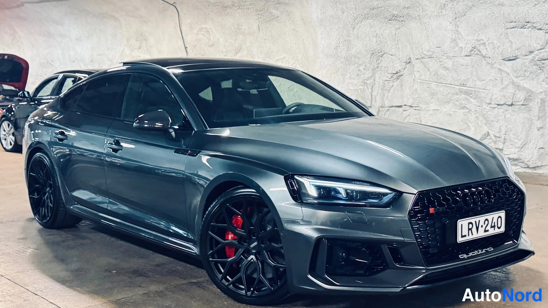 Etuviisto, nokka oikealle: Grey car angled to show the front left side. Black alloy wheels with red brake calipers are visible.
