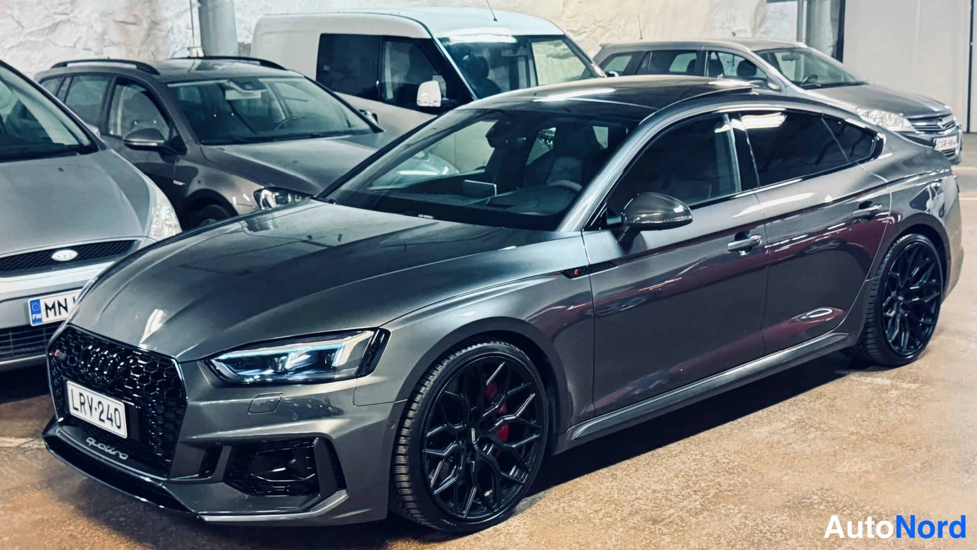 Etuviisto, nokka vasemmalle: Tummanharmaa Audi A5 Sportback sivulta vinosti edestä, mustat vanteet ja punaiset jarrusatulat, rekisteri.