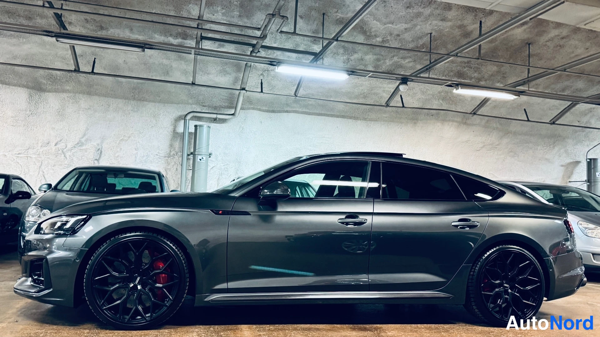 Etuviisto, nokka vasemmalle: Tummanharmaa Audi A5 Sportback:n koko sivuprofiili vasemmasta etukulmasta, mustat vanteet ja punaiset jarrusatulat.
