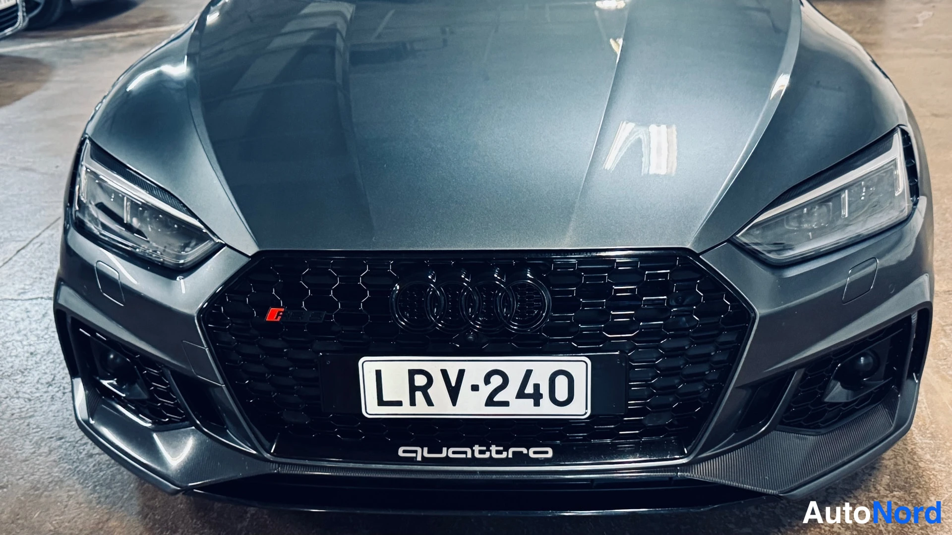Yksityiskohta: Close-up of the car's front face showing a black honeycomb grille, LED headlights, and a white license plate.