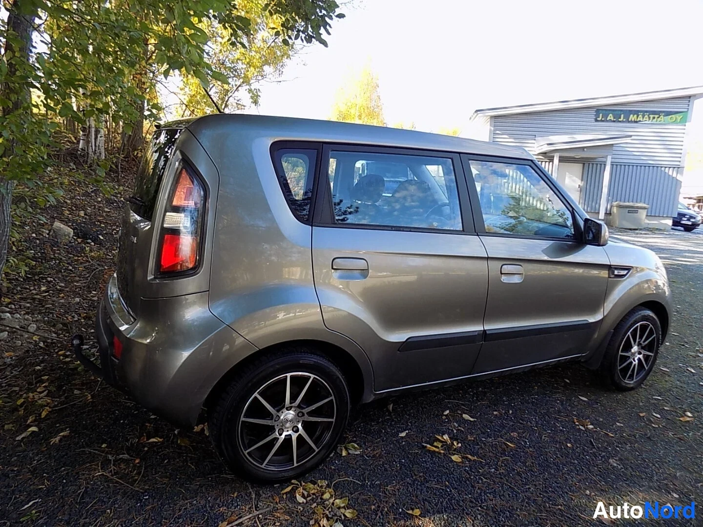 Takaviisto: Kia Soul -farmarimallisen viistoperän takakulma vasemmalta, harmaa runko ja takavalot näkyvissä puutarhataustassa.