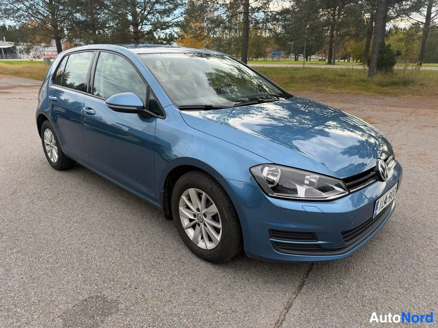 Etuviisto, nokka oikealle: Sininen henkilöauto kolmivartalokulmassa keulaa oikealle, etusäleikkö ja ajovalot näkyvät, asfalttialue ja puita taustalla.