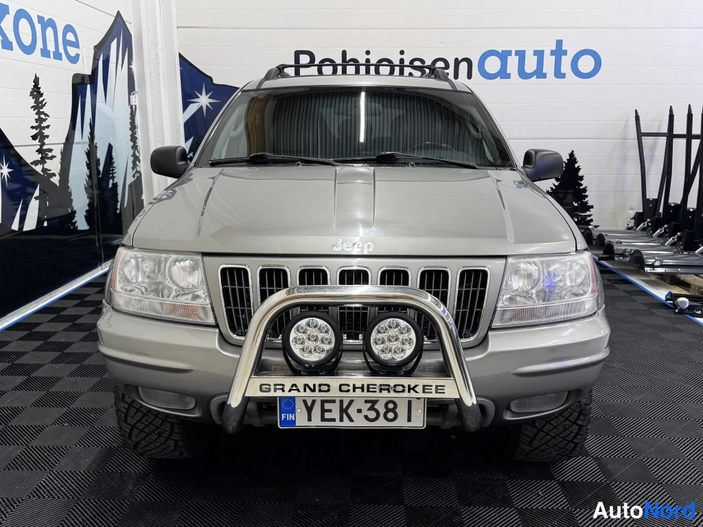 Edestä: Suora etunäkymä hopeanharmaasta Jeep Grand Cherokeesta; etusäleikkö, keulakaari kahdella lisävalolla ja rekisterikilpi.