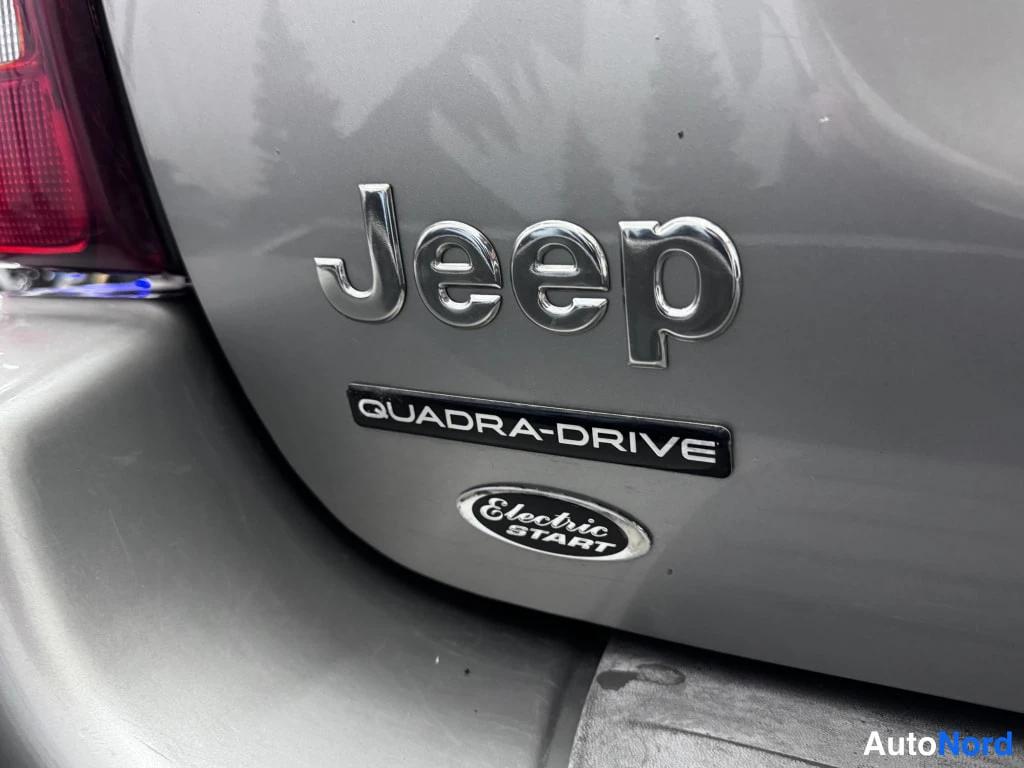 Muu: Lähikuva auton takapaneelista, jossa kromatut Jeep-merkki ja QUADRA-DRIVE-embleemi.