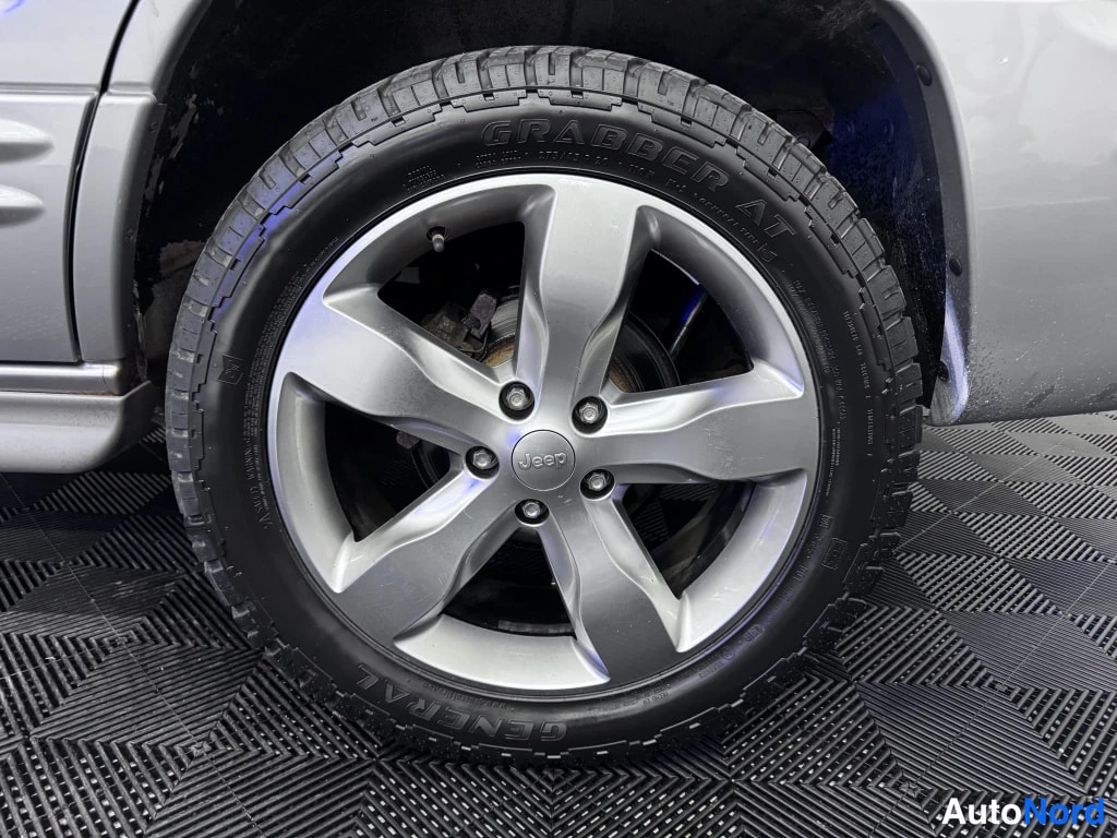 Yksityiskohta: Lähikuva auton pyörästä: hopeinen viisipuolainen Jeep-vanne ja General Grabber AT -kitkarengas, autopaikan ruudullinen mattolattia taustalla.